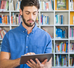 Ein junger Mann lernt am Tablet, er steht vor einem Bücherregal