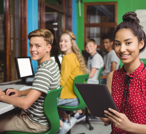 Ältere Schülerinnen und Schüler die jeweils an einem eigenen Computer sitzen. Eine Schülerin steht im Vordergrund, lächelt in die Kamera und hält ein Tablet in der Hand.