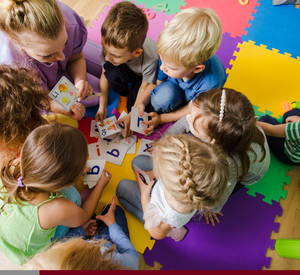 Symbolbild: Kitakinder mit einer Erzieherin auf dem Boden sitzend. Die Erzieherin zeigt den Kindern Buchstabenkarten.