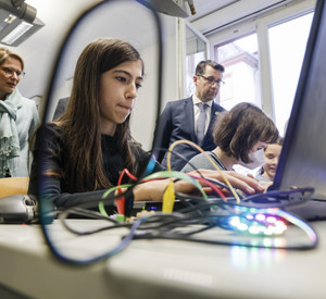 Eine Schülerin programmiert am Laptop eine Platine. Im Hintergrund steht Bildungsministerin Hubig und schaut der Schülerin zu. 