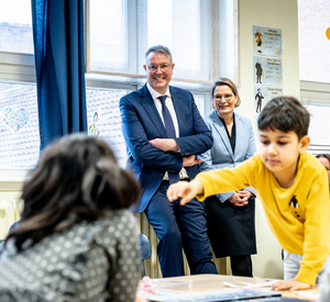 Ministerpräsident Alexander Schweitzer und Bildungsministerin Dr. Stefanie Hubig stehen im Hintergrund und beobachten die Arbeit der Lehrerin mit den Kindern an der Goethe-Schule.