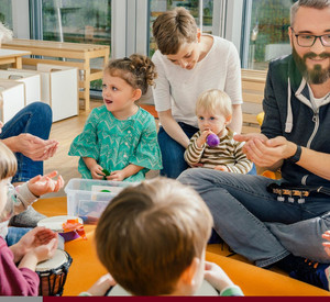Symbolbild: Kitakinder mit Erzieherinnen und Erziehern im Sitzkreis. Dabei musizieren die Kinder.
